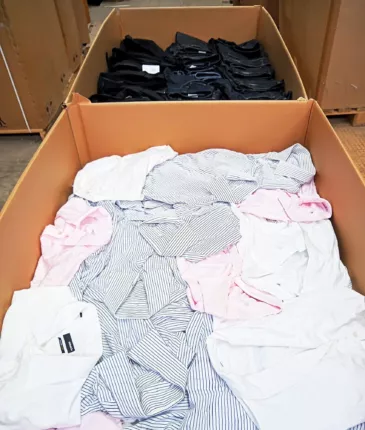 Großer Warenkarton voller Business-Hemden in Weiß, Rosa und blau gestreift, dahinter Jeans
