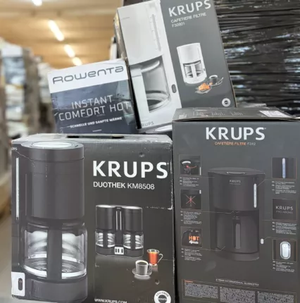 Nahaufnahme gestapelter Filterkaffeemaschinen und eines Heizlüfters vor Lagerpaletten