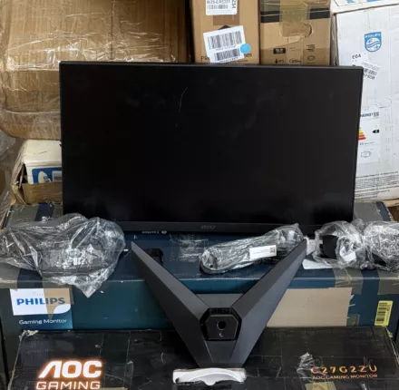 Ausgepackter Gaming-Monitor mit ausgeschaltetem Display, Standfuß und Zubehör vor gestapelten Monitor-Kartons