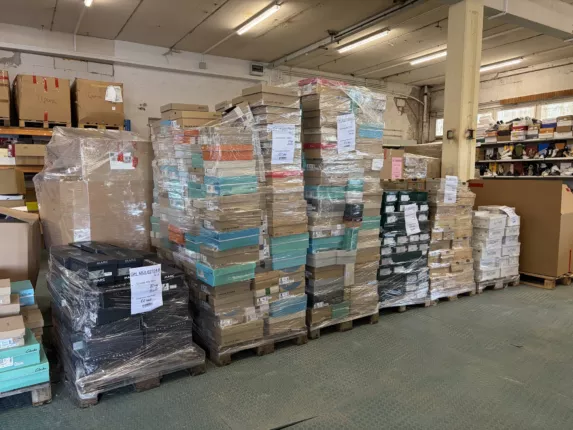 Lagerhalle mit mehreren folienverpackten Paletten voller Marken-Schuhkartons, im Hintergrund ein Regal mit einzelnen Schuhen